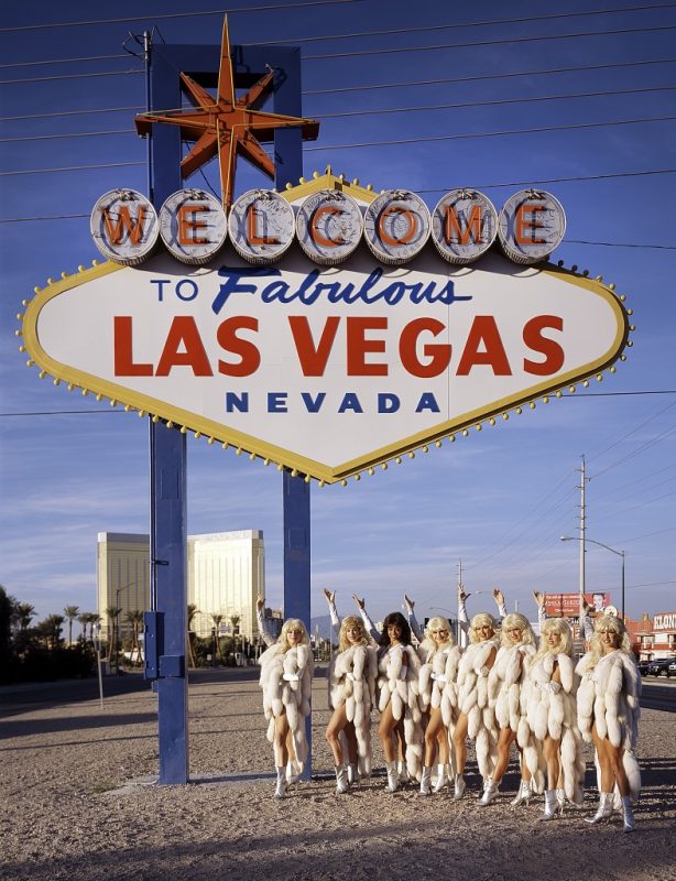 Eight women dressed in showgirl costumes stand under and gesture up to Las Vegas sign.