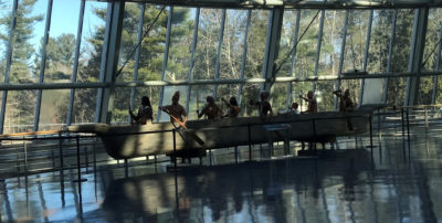 This photograph is of an exhibit of Native American figures in a canoe at the Mashantucket Pequot Museum & Research Center in Ledyard, CT. There are large windows in the background showing a wooded area outside.
