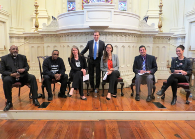 Six panelists seated and one standing on church alter at public plenary on Coltsville National Historical Park and gun violence. From left to right: Rev. Henry Brown, Warren Hardy, Sarah Pharaon, Hartford Mayor Luke Bronin, Thea Montañez, Iran Nazario, and Rebecca Stanfield McCown. Photo by Kelly Schmidt.