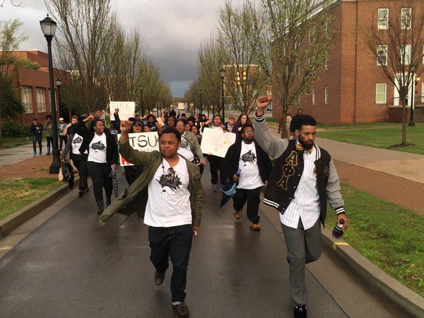 In this color photograph, students (mostly African American but also white) are pictured marching with signs in the street. Brick buildings and trees are in the background. The students are carrying signs (illegible).