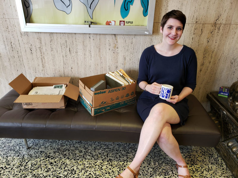 In this color photograph, a person sits on a brown tufted bench next to two cardboard boxes that contain books and papers. The person has short brown hair and is wearing a navy blue dress. They are holding a blue and white mug with the NCPH logo on it while smiling.