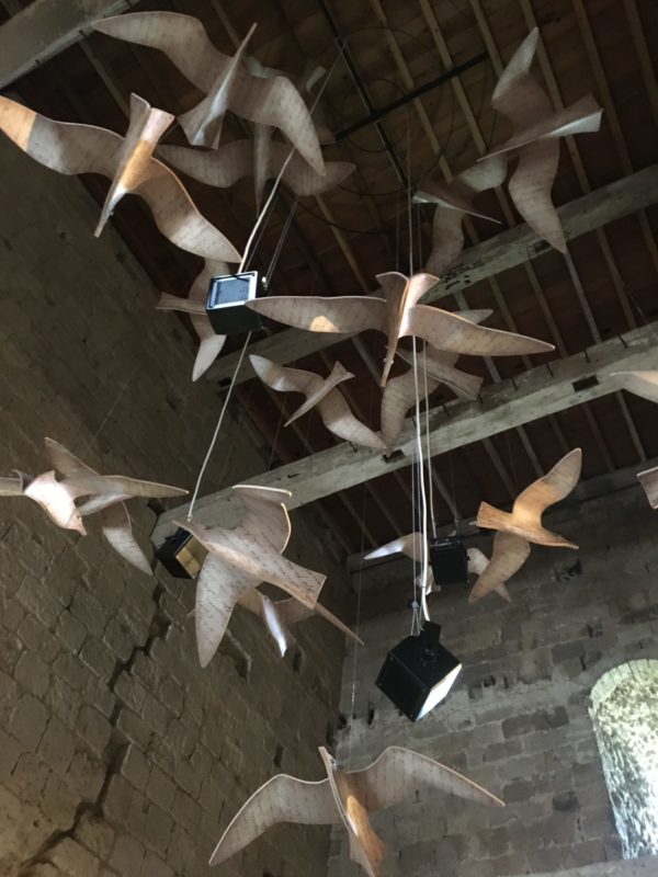 This is a color photography of artistically, three-dimensional renderings of seagulls having from a ceiling by string. There are black speakers interspersed among the seagulls.