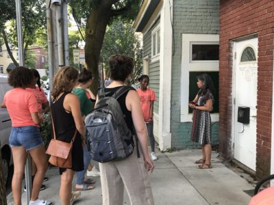 This is a color photograph of a group of people on a sidewalk taking a walking tour. The people are looking at a tour leader and brick building. One of the brick buildings is painted light green.