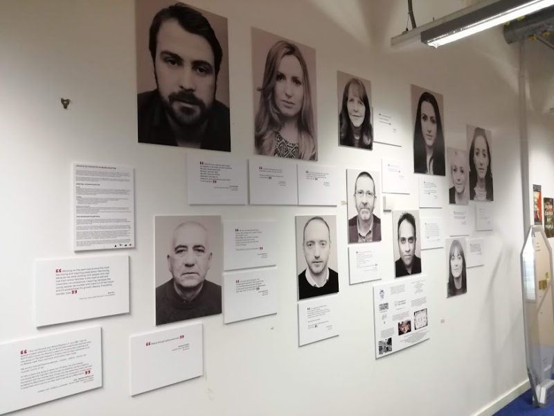 This is a color photograph of a wall covered with black and white photographs of people's faces, quotations, and exhibit labels.