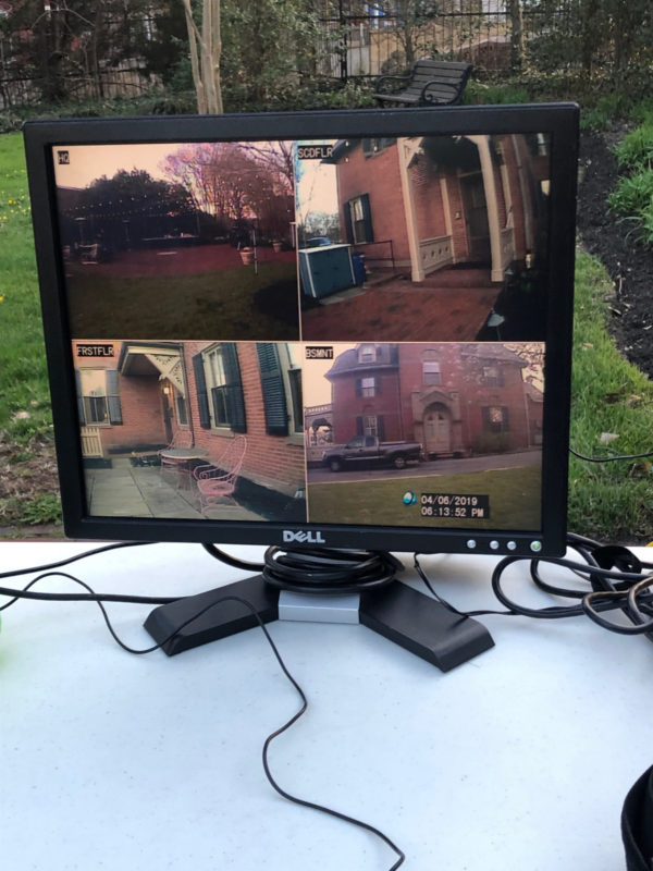 This is a color photograph of a computer screen on a table outside. The computer screen is showing camera feeds from different locations outside the house pictured above.