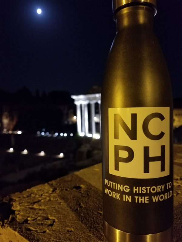 The image is taken at night. A NCPH water bottle on a wall-like surface is in the foreground. In the background is an illuminated building and a full moon.