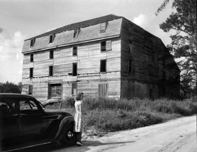 In the foreground, a woman stands in front of a car. A four story wooden barn is in the background.