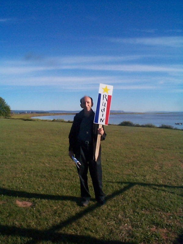 This is a color photograph of a man standing on grass with a body of water in the background. There are very few clouds in the sky (which is blue). The May is wearing dark pants and jacket as well as a blue shirt. He is carrying a narrow sign on a wooden post. The sign reads "RUDIN" down the middle in blue. There is a yellow star at the top. On the left, there is a blue stripe. There is a red stripe on the right.