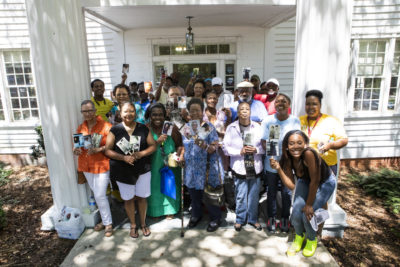 This is a photograph of a group of about two dozen African American individuals of all ages standing in front of a White House holding up photographs.