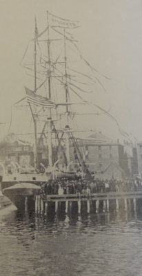 This is a black and white photograph depicting a docked sailing vessel flying the American flag and surrounded by a crown of people.