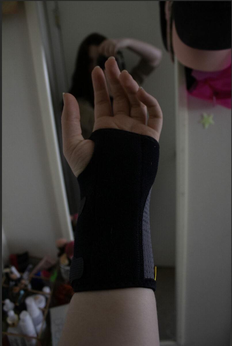 This is a color photograph of a person's reflection in a mirror (background) taking a photograph of their left wrist and hand in a black brace (foreground).