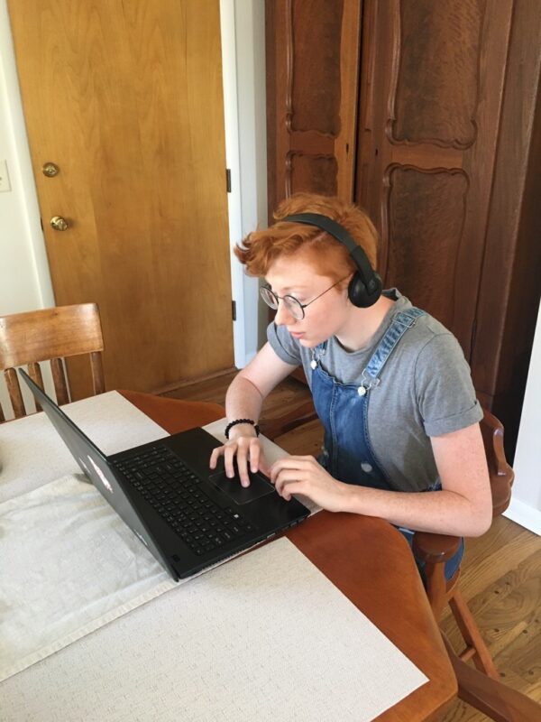 This is a color photograph of a person in their twenties sitting down in front of a laptop. They have red hair and a re wearing glasses, a headset, and overalls.