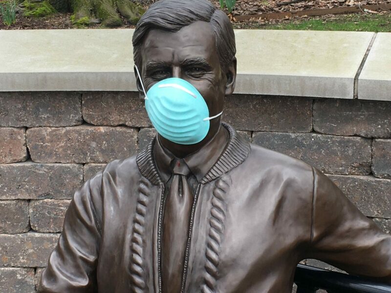 This is a color photograph of the upper portion of a sculpture of Fred Rogers, seated, and wearing a mask.