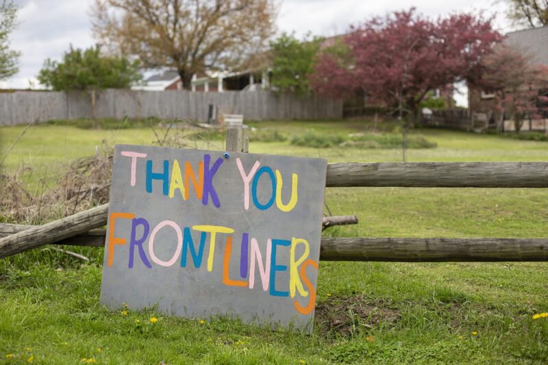This is a color photograph of a homemade sign reading "THANK YOU FRONTLINERS." It is attached to a fence in a field.
