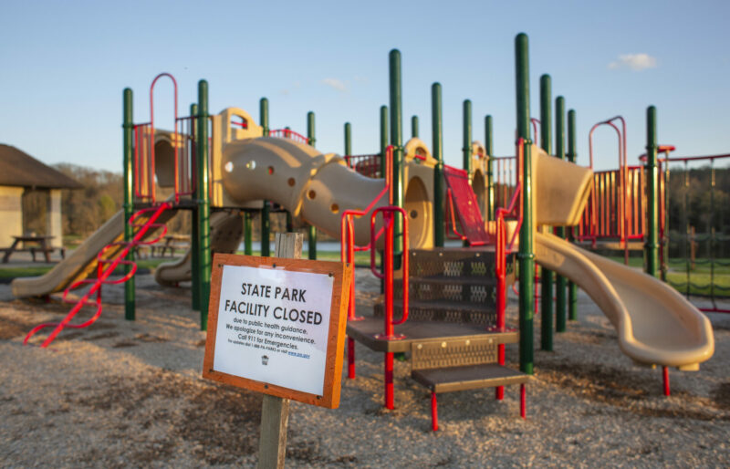 This is a color photograph of an empty playground. There is a play set in the background and a closure sign about COVID-19 in the foreground.