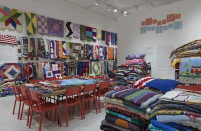 A full gallery with colorful knitted blankets covering the walls, tables, and piled high all over the room, and a sign saying “Welcome Blanket