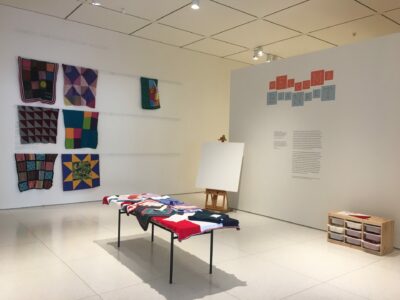 A mostly empty gallery with a few colorful knitted blankets and a sign saying “Welcome Blanket”