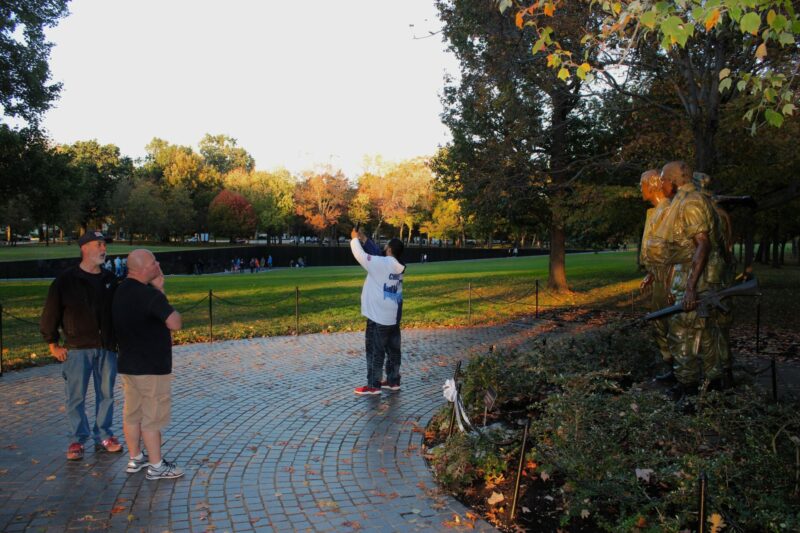 Three men look at "The Three Servicemen" sculpture. One man is taking a selfie.