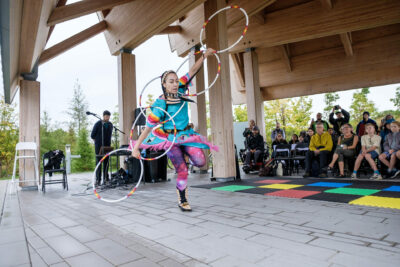 A woman performs a hop dance under a shelter while the audience looks on. 
