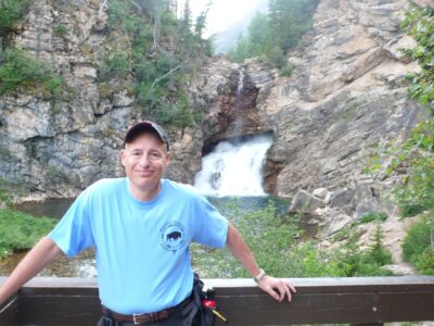 A smiling man with light-tan skin, wearing a blue shirt and a baseball cap stands on a platform overlooking a waterfall