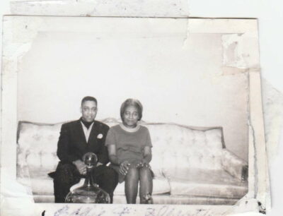A Black couple wearing formal dress are seated on a white upholstered couch
