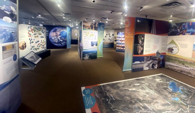 This photo depicts an exhibition with no visitors in it. There are large panels and screens along three walls, and a table-height map in the foreground.