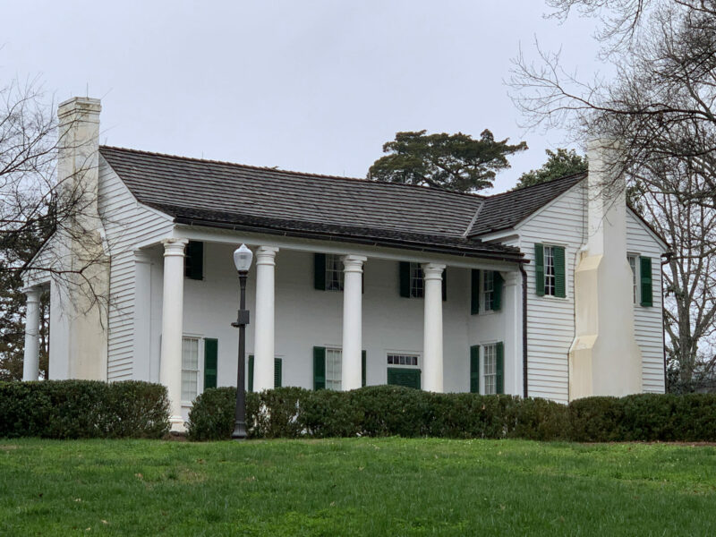 A large house with two chimneys, white-painted siding, green-painted shutters, and four Corinthian columns stands on a green field surrounded by bushes. 