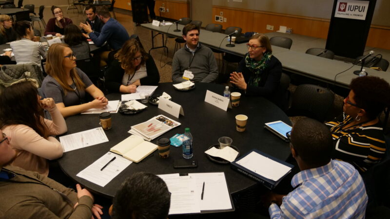 Seven students sit at a round table with notebooks out listening to a museum professional discuss her career in the field.