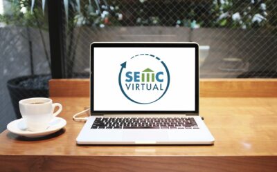 A cup of coffee or tea sits on a saucer next to a laptop displaying a circular logo and the text “SEMC Virtual” on the screen. The photo is taken from the perspective of the computer user, who is sitting inside at a wooden table and can see plants outside through a large window.