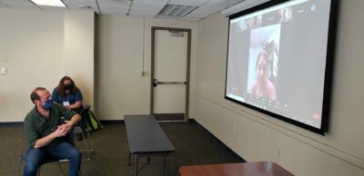 Two men wearing face coverings and sitting in a meeting room watch a screen where a woman speaks via Zoom conferencing software.