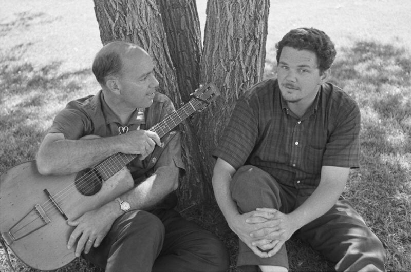 Sam Hinton and Barry Olivier sit in front of a tree. Hinton, with guitar, looks over at Olivier.