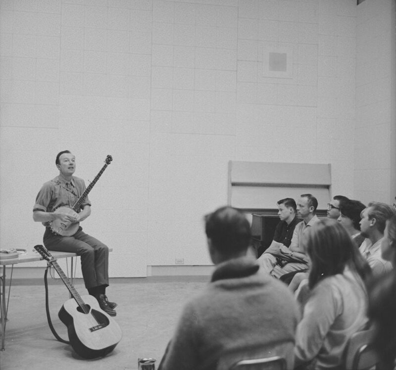Pete Seeger sits with banjo at the front of a classroom while audience listens.