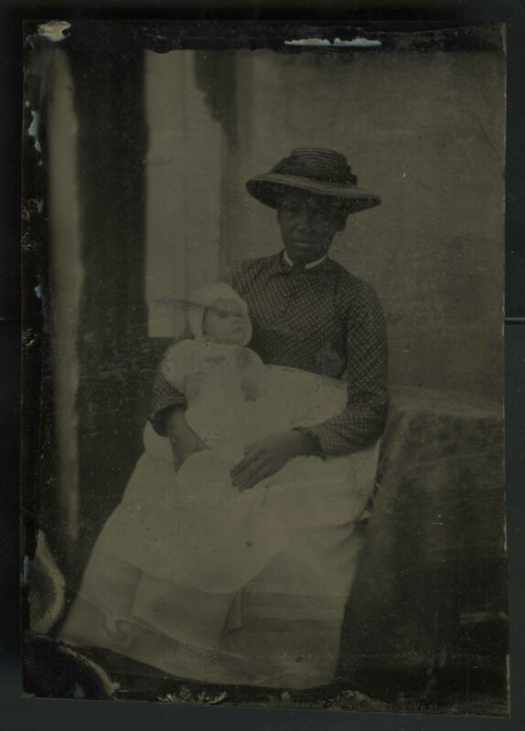 A photographic tintype of an unidentified Black woman holding an unidentified white child. The woman is in a floor-length polka dot dress and a straw hat. She is also wearing a white apron. She holds a white infant wrapped in a white blanket that looks up at her. The woman is looking right at the camera with an emotionless expression, and she could be anywhere from 20 to 35 years old. They sit together on a chair indoors, next to a small table and sunlight coming in from the window. 