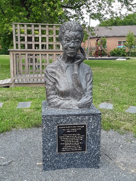 Stone bust of Black writer Gwendolyn Brooks. The bust is situated on a marble platform with a black plaque with lettering. There is grass and a playground in the background.