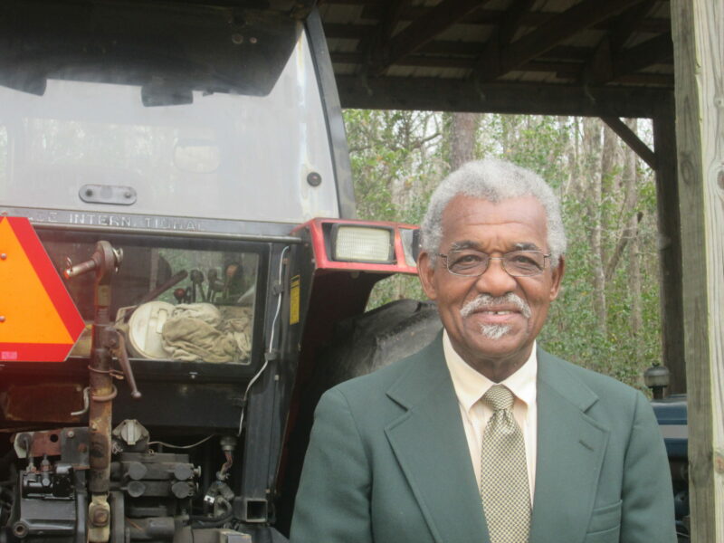 An older Black man smiles into the camera. He wears glasses and his hair is mainly silver. He has a mustache and tiny goatee. He is wearing a olive gray suit jacket, a light yellow dress shirt, and gold checked tie. There is a large piece of farm equipment behind him.
