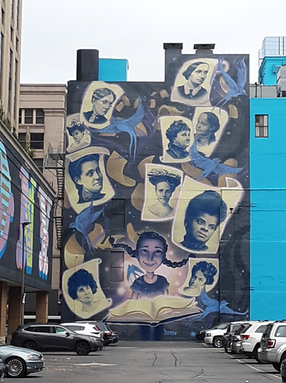 Mural of several historical female suffragists on a wall overlooking a parking lot in Chicago. Their images are imposed in individual portraits along a black background with blue birds flying throughout. The mural is located between two buildings. 