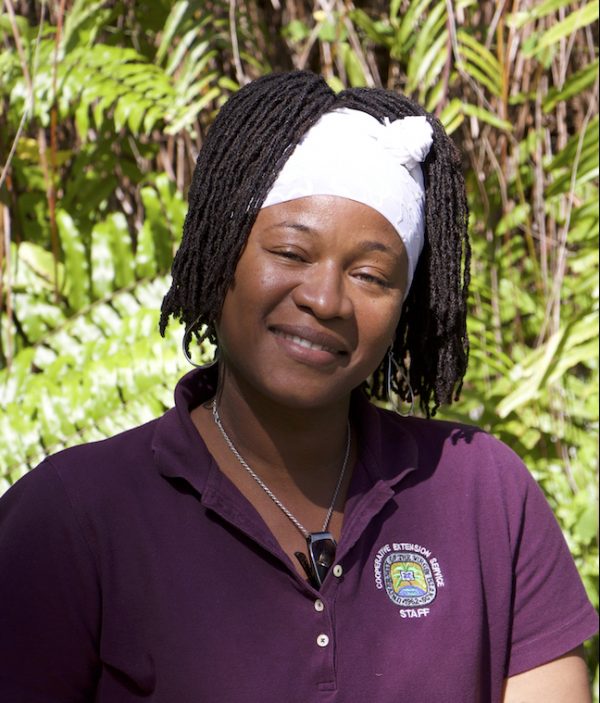 A Black woman smiles, looking into the camera. She is wearing a purple polo shirt, with a logo on the right side. Here hair is in small locs and held off her face with a white cloth.