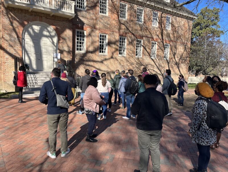 The image shows a large group of individuals outside of a large brick building, known as the Wren or College Building . The sky is very blue and there are tree shadows across the brick building. Several individuals from the group are preparing to walk up the stairs into the building’s cream-colored doors.