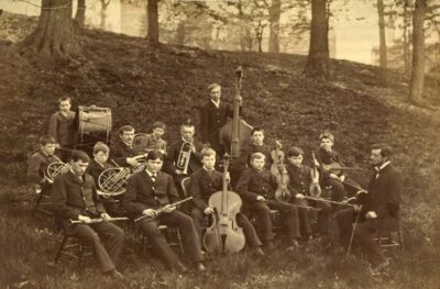 In black and white, a group of boys and young men holding musical instruments are seated on a slope in the woods. A leader with a violin bow faces the band. Members hold violins, clarinets, horns, and a cello. A boy at the back holds a large drum.