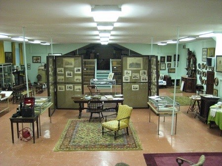 A large, open room with mint green walls is filled with a range of exhibits. In the center of a room, a rug, chairs, and tables recreate a nineteenth-century administrator’s desk surrounded by glass cases with historical objects in them. Behind the desk, photographs appear in standing display cases. In the back of the room is a set of stairs surrounded by bookcases.