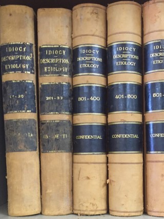 Five beige, leather-bound books sit next to each other. Their spines read, “Idiocy Descriptions Etiology” followed by case numbers and “Confidential.”
