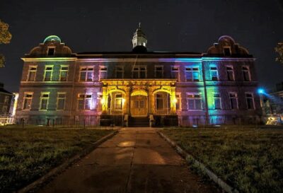 A three-story brick building at night. The front of the building is illuminated with colored lights. 