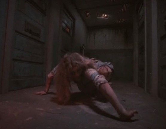 A young white woman with long hair wearing old clothes crawls along the floor of a dark building. Her wrists and elbows are twisted as she moves.