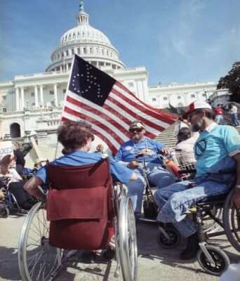 A group of disabled protesters in wheelchairs with a universal access flag in front of the United States Capitol.