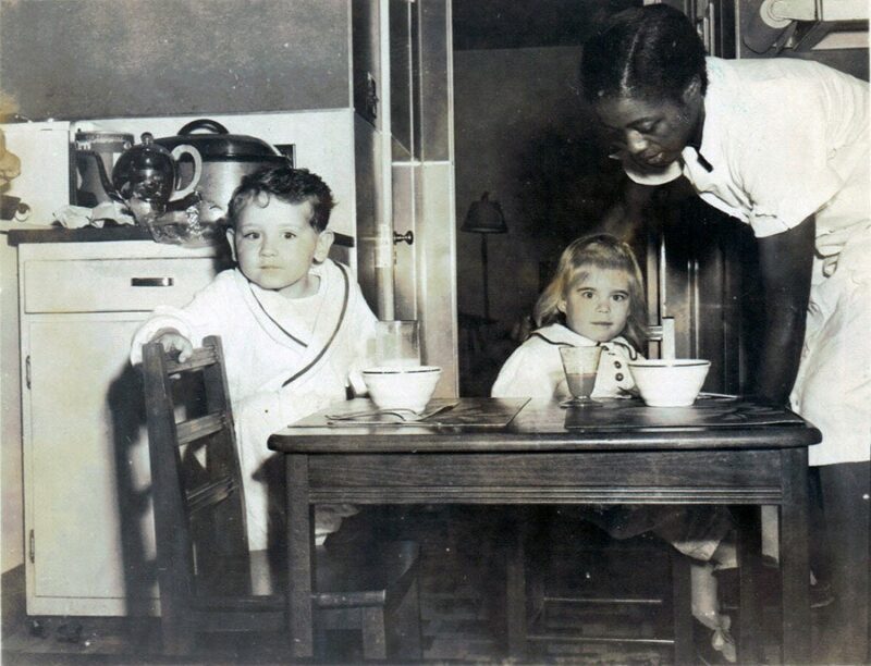 Two young white children, one standing and one sitting, are seen at a child-sized table in a kitchen. A Black woman in a white dress leans over to help the girl.