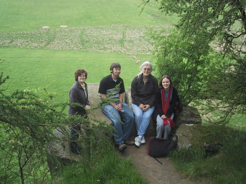 Color photo of four people sitting on large boulders with green grass in the background. 