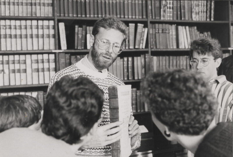Black and white photo of four people facing Ed King, who is holding a book and standing in front of large book shelves.