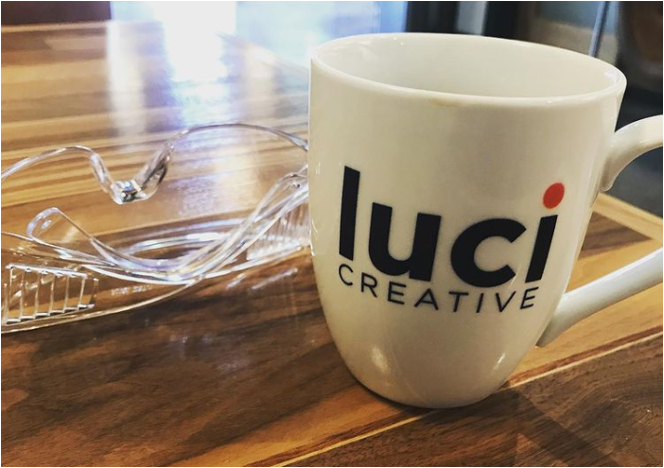 A closeup shot of a white mug with the Luci Creative logo and a clear set of safety glasses on a wooden table