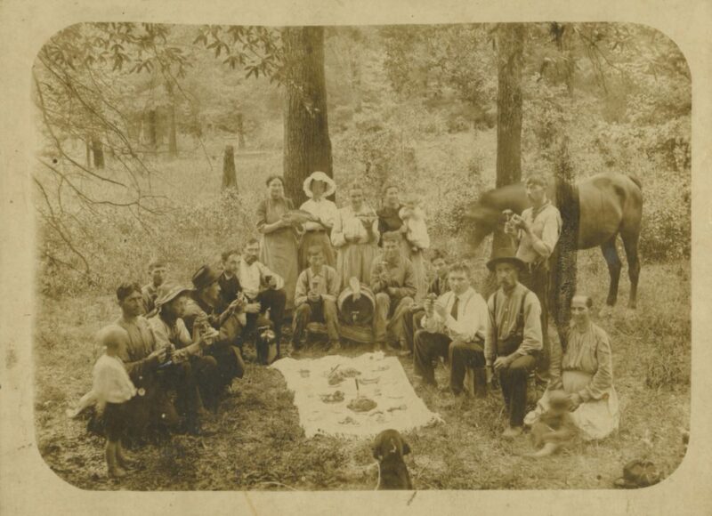 A group of eighteen people gathered around a picnic blanket with food on it posing for a photograph with a horse and woods in the background and a dog in the foreground