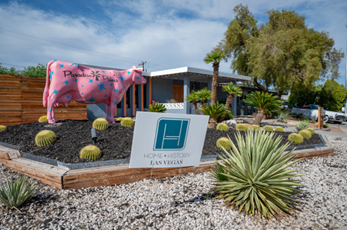 A model of a pink cow located outside on top of mulch and other outdoor greenery adjacent to a building in the Paradise Palms Historic Neighborhood. The sky is blue and filled with clouds. There is a tree outside of a home.